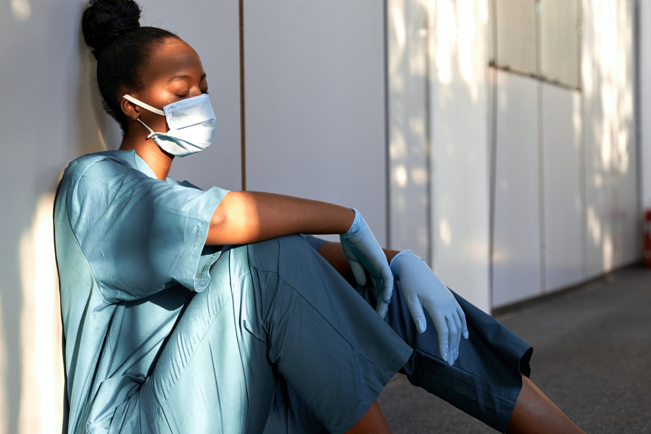 Tired exhausted female african scrub nurse wears face mask blue uniform gloves sits on hospital floor. Depressed sad black ethic doctor feels fatigue burnout stress, lack of sleep, napping at work.