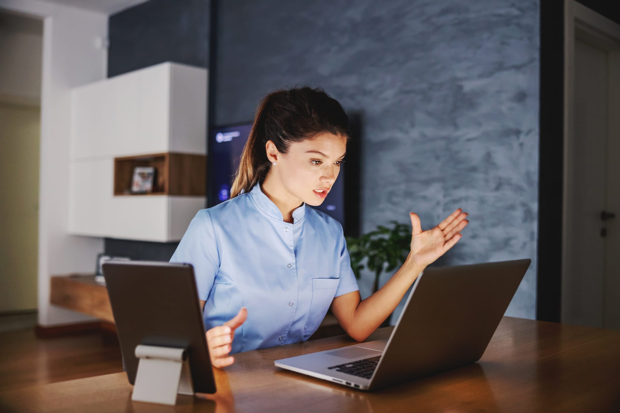 Nurse sitting at home and giving online advices. On table there are tablet and laptop.